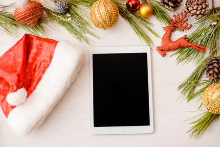 Happy Christmas background. Top view on tablet computer with toys and Santa Claus hat on wooden table. Modern festive decoration ready for joyful message or shopping timeの写真素材