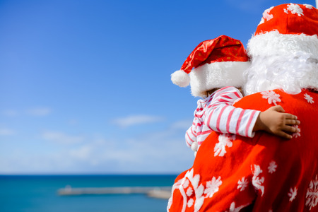 Happy Merry Christmas. Child hugging Santa Claus father together wearing red hats over sunny blue sky background outdoors. Back viewの写真素材