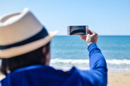 Back view shot of man holding smartphone photographing selfie or beach background outdoorsの写真素材