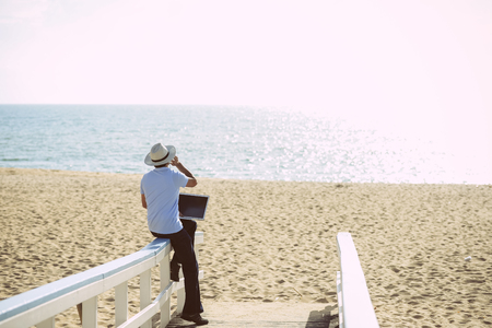 Busy male working on the laptop and talking on mobile. Man in hat speaking on smartphone over blue sky and ocean beach backgroundの写真素材