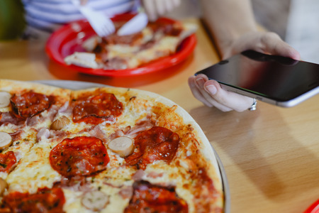Closeup of people eat and work. Hands, sliced pizza and using smartphone on restaurant table background. Communication, friendship leisure lifestyleの写真素材