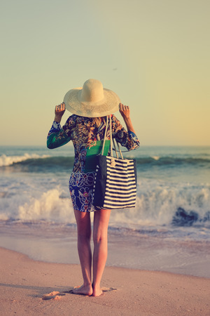 Holiday lifestyle woman in straw hat on a beach holding bag. Recreational enjoyment walk along sunny outdoors background coastline. Idyllic remote relaxation seashore landscapeの写真素材