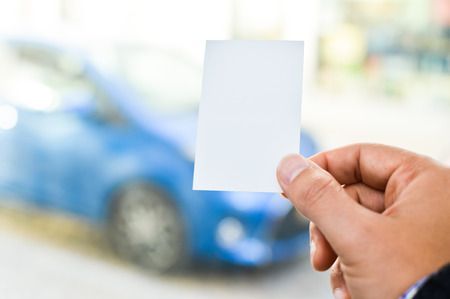 Close up on hand of a businessman holding white business card over blurry car office background. Professional person showing introductionの写真素材