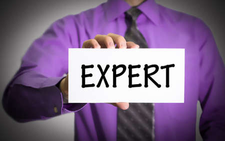 Businessman in blue shirt and gray tie shows a card with the inscription expert. Man on a gray background. Selective focus.の写真素材
