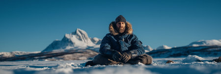 Man in winter gear sitting on snow looking at snowy mountains under clear sky. Adventurer man...