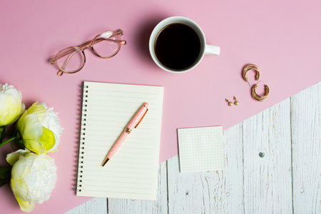 Women working items with white roses, notebook, coffee cup and earrings over pink and wooden table.