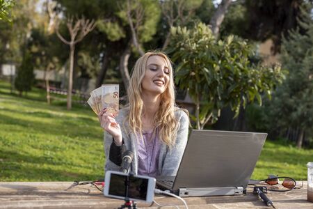 Female blond blogger demonstrates the money earned from internet services - working outdoors -  cash...