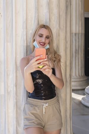 beautifil blond girl smiling with face mask after lockdown reopening - She is enjoying her walk having fun, chating with friends, taking selfies outside marble building - socializing again projectの写真素材