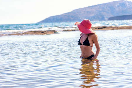 rear view of a sexy mature woman in black bikini and red hat walking at the sand of a tropical beachの写真素材