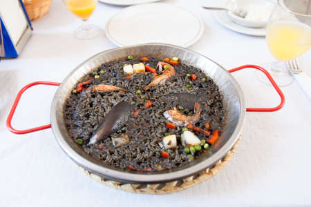 Paella negra on the pan, white table and glasses with juiceの写真素材