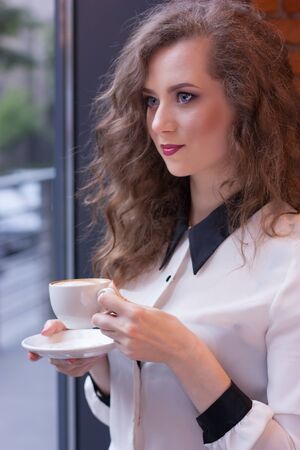 Beautiful girl with coffee in a white shirt at the windowの写真素材
