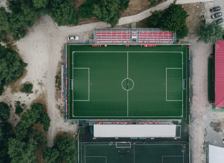 Top-down drone perspective of a green soccer field with red stadium seats, surrounded by trees and pathways. Professional sports complex architecture.