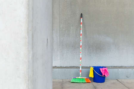 Colorful cleaning equipment (broom, bucket, rag and gloves) in front of a gray, monotonous concrete...