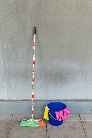 Colorful cleaning equipment (broom, bucket, rag and gloves) in front of a gray, monotonous concrete...