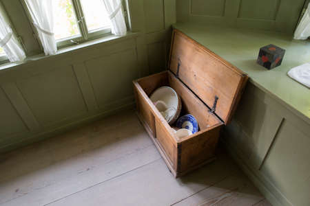 An open brown wooden chest filled with dishes, standing in a room with light green walls.