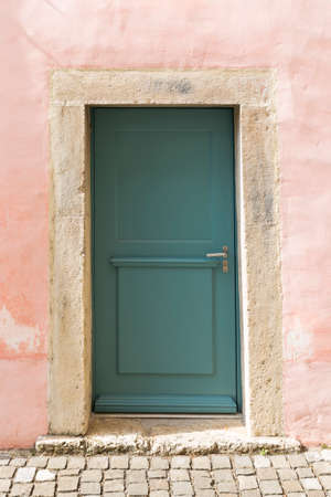 A green painted wooden door in a massive, pink painted stone wall with solid stone frame and cobble stone pavement.の写真素材