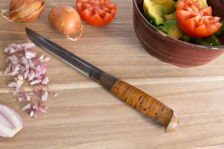 Close view on a brown wooden cutting board with a big knife, a red bowl with avocado pieces, two...