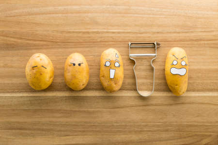 Fresh potatoes with cartoon style faces laying on a brown cutting board and scared to death from a peelerの写真素材