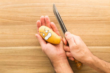 a fresh potato with cartoon style face scared to death while being held in one hand. The other hand holds a big knifeの写真素材