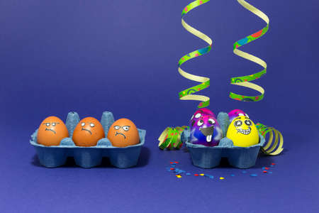 Group of colorful painted Easter eggs with funny cartoon style faces, confetti and paper streamers and group of grumpy looking brown eggs in light blue egg boxes on purple backgroundの写真素材