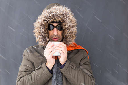 Portrait of a single male winter adventurer wearing a warm green coat with fur hood, an orange scarf and black retro style goggles drinking from a metal cupの写真素材
