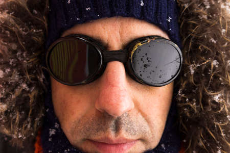 Portrait of a single male winter adventurer wearing a warm green coat with fur hood, a blue ski cap, an orange scarf and black retro style gogglesの写真素材