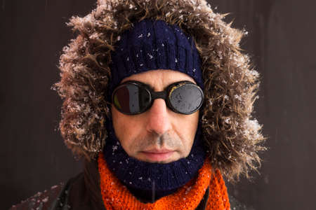 Portrait of a single male winter adventurer wearing a warm green coat with fur hood, a blue ski cap, an orange scarf and black retro style gogglesの写真素材