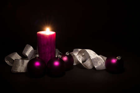 Arrangement of beautiful illuminated purple colored Christmas balls with a silver and white colored ornate ribbon and a burning candle on black backgroundの写真素材