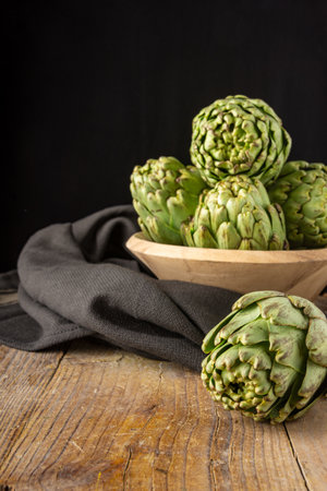 Top view of green artichokes in wooden bowl with black cloth, with selective focus, on wooden table, black background, vertical, with copy spaceの写真素材
