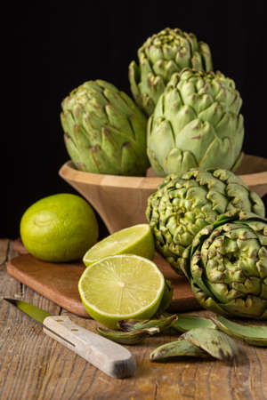 View of green artichokes in bowl, limes on table and knife, on wooden table, with selective focus, black background, verticalの写真素材