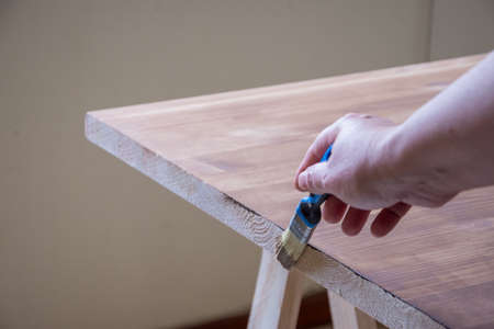 Close-up of woman's hand with brush going over the paint on a pine wood table, horizontal
