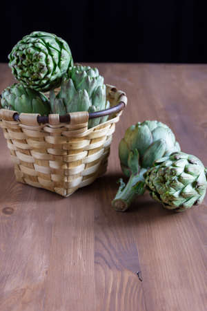 Top view of rustic basket with artichokes on wooden table with two artichokes, black background, verticalの写真素材