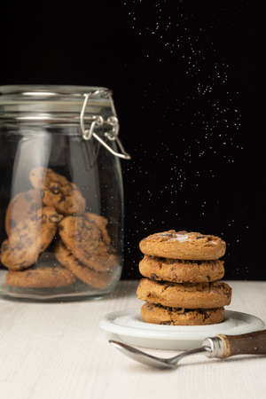 View of stack of chocolate cookies on plate, with falling sugar, spoon and glass jar with cookies, selective focus, on white wooden table, black background, verticalの写真素材