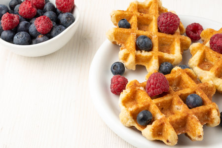 Aerial view of white plate with waffles with raspberries and blueberries, bowl with raspberries and blueberries, selective focus, on white table, horizontalの写真素材
