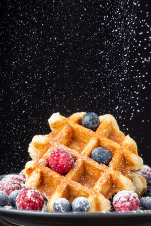 Close-up of waffle with raspberries and blueberries on dark plate, with sugar falling, black background, verticalの写真素材