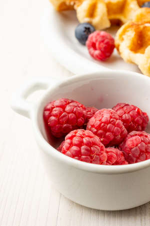 Top view of bowl with raspberries, plate with waffles with raspberries and blueberries on white plate, selective focus, verticalの写真素材