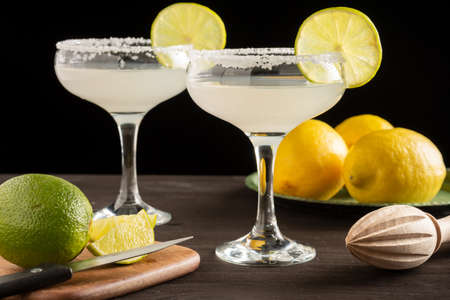 Close-up of two glasses with margarita cocktail, on dark wooden table with limes and lemons, black background, horizontalの写真素材