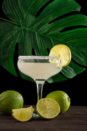 View of limes and glass of margarita cocktail, selective focus, on wooden table, black background with green leaf, verticalの写真素材
