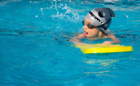 A young boy swimming with a floating yellow board  in a blue water of swimming pool