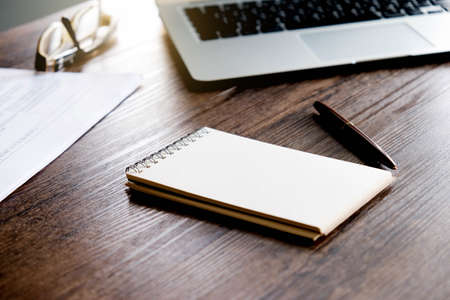 Office workspace with blank notebook , pen , paper sheet and laptop on the wooden table with warm fall colors and soft-focus.の写真素材