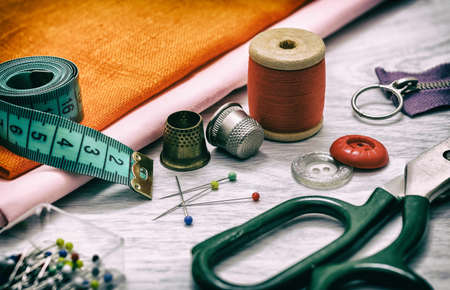 Set of sewing accessories. Sewing accessories for sewing: orange cloth, thimbles, spools of thread, tailor's scissors, a set of pins and a tape measure on a gray wooden table. retro styleの写真素材