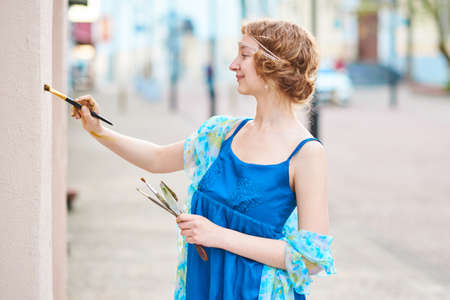 Beautiful girl-artist on the street in blue dress, draws on the wall for any purposeの写真素材