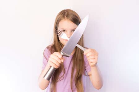 Little cute girl holding a knife and fork crosswise on a light background for any purposeの写真素材