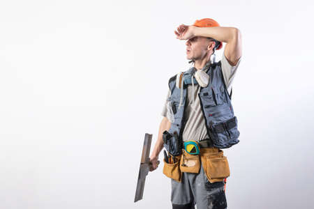 Tired builder with a spatula. In work clothes and hard hat. On a light gray background.