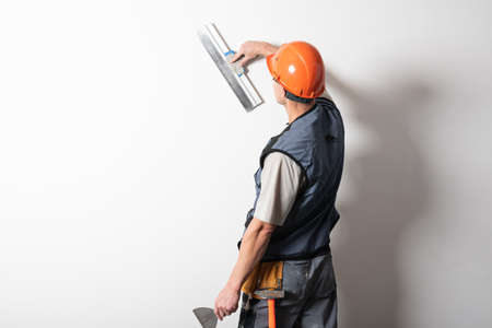 The builder smoothes the wall with a spatula. In work clothes and hard hat. On a light gray background..の写真素材