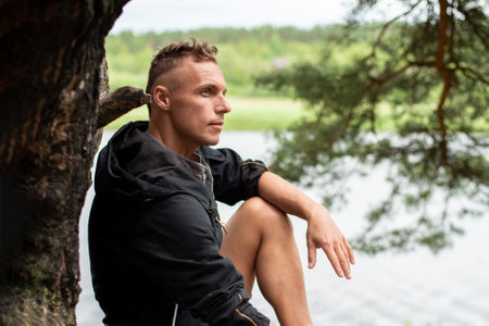 A man on the shore of a lake, sitting on the roots of a tree. For any purpose.の写真素材