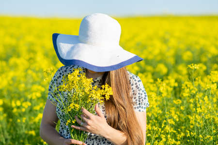Girl in a rapeseed field, in a hat, with a bouquet of rapeseed in her hands. High quality photo