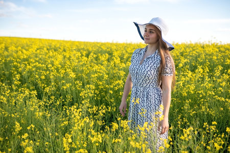 Beautiful girl in a hat and dress, in a rapeseed field, against the sky. High quality photo