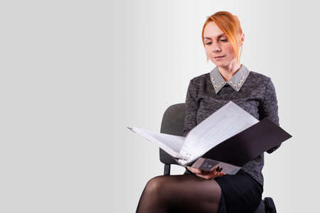 Business woman sitting on a chair with an open folder in her hands, on a gray background. High...