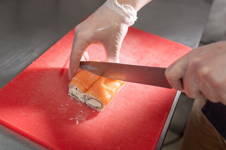 The cook cuts sushi roll with salmon, cheese and cucumber on a red board. high quality photoの写真素材